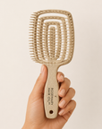 Hand holding a beige hairbrush with 'Rosemary Hair Oil Co.' branding on a beige background, Beige hairbrush with 'Rosemary Hair Oil Co.' branding on a beige background, detangle brush, tangle teaser, flexi brush