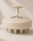 A speckled beige hair massager with a handle on top and multiple short spikes around the base, shown on a warm beige background. The lid is printed with the brand name 'ROSEMARY HAIR OIL co. Rosemary hair oil, scalp massager, soothe, essential, hair growth, stimulate, health, haircare, UK