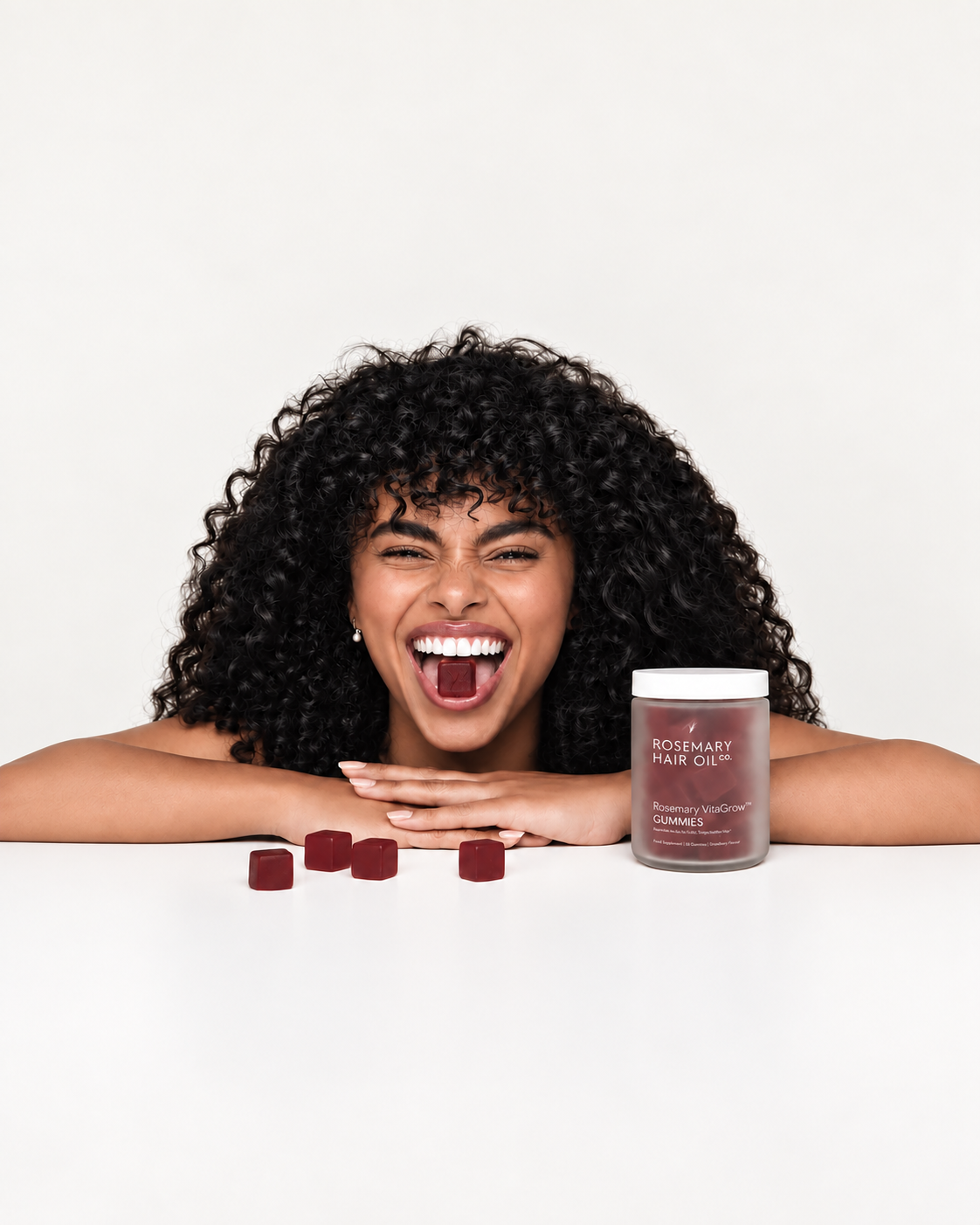 A smiling woman with voluminous curly black hair leans forward over a white surface, with several dark red gummies in front of her and a jar of Rosemary Hair Oil Gummies to the right. The background is a clean white studio backdrop.