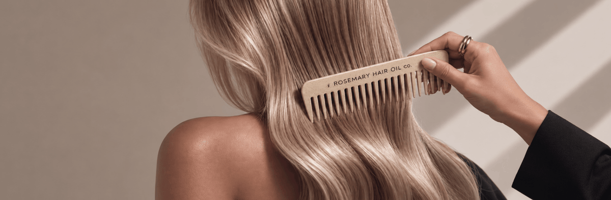A close-up of a person with long, wavy blonde hair being combed with a light wood comb labeled 'ROSEMARY HAIR OIL'. Rhoco Ritual
