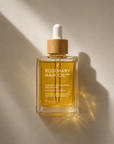 A clear glass dropper bottle of Rosemary Hair Oil Co. rosemary hair oil intense growth drop, filled with golden yellow oil, set against a warm beige background with dramatic diagonal sunlight and shadows.