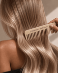 A close-up lifestyle shot of a person with long, wavy blonde hair using a light beige wooden comb branded '