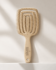 A natural hairbrush with a rectangular head and a long handle, standing upright on a white surface against a light beige background with soft diagonal shadows. The handle is printed with 'ROSEMARY HAIR OIL CO. Beige hairbrush with 'Rosemary Hair Oil Co.' branding on a beige background, detangle brush, tangle teaser, flexi brush