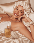 A woman in a satin shower cap and white robe reclines on a satin bed with a bottle of Rosemary Hair Oil and a scalp massager in the foreground. Rosemary hair oil with a dropper and two satin hair bonnets on a light background. Award-winning Hair Growth Starter Kit with Rosemary Hair Oil Intense Growth Syrup. RHOCo. HairHalo | Pure Silk Bonnet. Scalp Soothe Essential Massager
